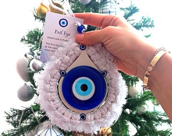 May include: White crocheted ornament with a blue and white evil eye charm. The ornament is hanging on a Christmas tree. The text on the tag reads "Evil Eye Good Luck & Protection The Evil Eye symbol protects from negativity".