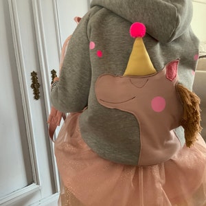 Könnte beinhalten: Ein grauer Kapuzenpullover mit einem rosa Einhorn-Applikationen auf der Rückseite. Das Einhorn trägt einen gelben Partyhut mit einem rosa Bommel oben. Der Pullover hat rosa Punkte auf den Ärmeln. Das Mädchen trägt einen rosa Tüllrock mit Glitzer.