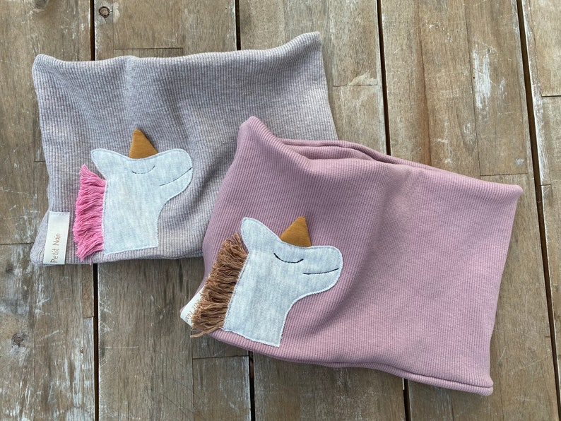 May include: Two ribbed knit neck warmers, one gray and one mauve, each adorned with a unicorn applique. The unicorns have a gray face, a gold horn, and a pink or brown mane. The gray warmer has a small label that says "Petit Nie".