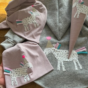 Könnte beinhalten: Ein Set aus drei rosa und grauen Baby-Hoodies mit Einhorn-Motiv. Die Outfits beinhalten eine Kapuzenjacke, eine Mütze und ein Lätzchen. Jedes Teil zeigt ein schwarz-weiß geflecktes Einhorn mit einem rosa-blauen Partyhut.