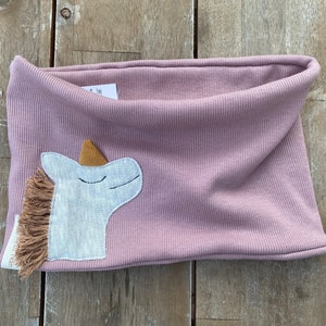 May include: A dusty rose-colored neck warmer with a ribbed texture. A white unicorn applique with a brown mane and horn is sewn on the front. The neck warmer is made of soft fabric and is designed for children.