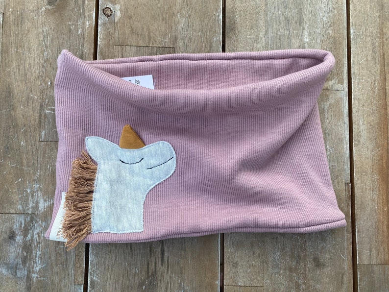 May include: A dusty rose-colored neck warmer with a ribbed texture. A white unicorn applique with a brown mane and horn is sewn on the front. The neck warmer is made of soft fabric and is designed for children.