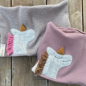 May include: Two ribbed knit neck warmers, one gray and one mauve, each adorned with a unicorn applique. The unicorns have light blue faces, brown horns, and pink and brown manes. The label "Petit Nain" is visible on each warmer.