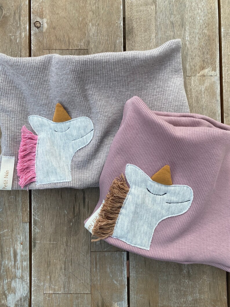 May include: Two ribbed knit neck warmers, one gray and one mauve, each adorned with a unicorn applique. The unicorns have light blue faces, brown horns, and pink and brown manes. The label "Petit Nain" is visible on each warmer.