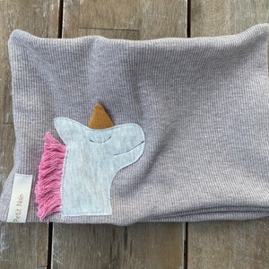 May include: A light gray ribbed knit neck warmer with a unicorn applique. The unicorn has a white head, a gold horn, and a pink fringe mane. A small tag with the text "Petit Nain" is sewn on the side.
