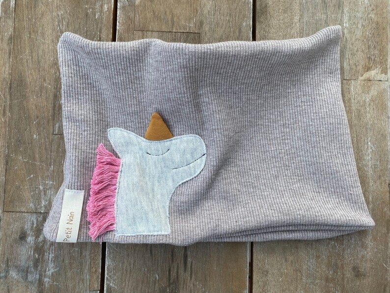 May include: A light gray ribbed knit neck warmer with a unicorn applique. The unicorn has a white head, a gold horn, and a pink fringe mane. A small tag with the text "Petit Nain" is sewn on the side.