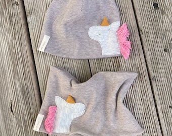 May include: A gray knit hat and matching neck warmer with a unicorn applique. The unicorn has a pink mane and a gold horn.