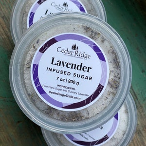 May include: Two clear plastic containers filled with white sugar infused with lavender. The label reads "Cedar Ridge Lavender Infused Sugar 7 oz | 200 g. Ingredients: Pure Cane Sugar and Culinary Lavender. CedarRidgeTrails.com"