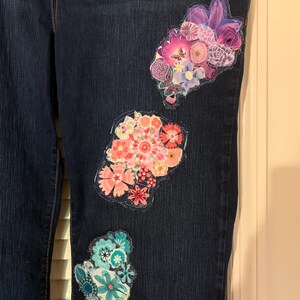 May include: Dark blue denim jeans with three floral patches in shades of pink, purple, and teal. The patches are made of various floral fabrics and are sewn onto the jeans. The jeans have a classic five-pocket design.