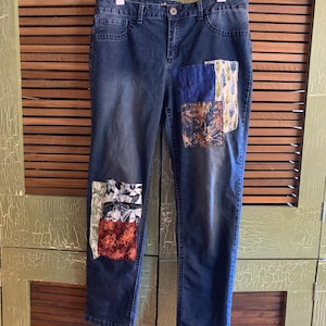 May include: Blue denim jeans with patchwork details on the knees and right thigh. The patches are made of various fabrics in shades of blue, green, orange, and white.
