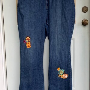 May include: Dark wash denim jeans with embroidered floral patches on the knees. The jeans have a flared leg and a classic five-pocket design. The floral patches feature orange, yellow, and teal colors.