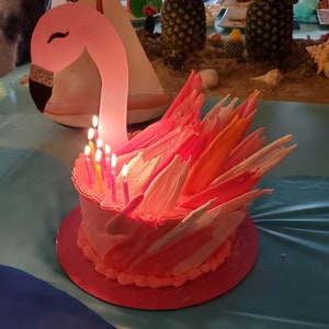 May include: A pink flamingo-shaped cake with lit candles. The cake has a pink base and is decorated with pink, orange, and white accents. The cake sits on a red plate, with a flamingo head and neck rising above it.
