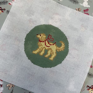 May include: A needlepoint canvas with a golden retriever design inside a green circle. The dog has a red harness, set against a white mesh background. The canvas is likely for a festive project, possibly Christmas-themed.