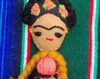 May include: A handmade felt doll of Frida Kahlo with black hair, a pink dress, and a yellow top. The doll is wearing a flower crown and has a painted unibrow.