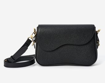 Puede incluir: Bolso bandolera negro con solapa curva y correa ajustable. El bolso presenta herrajes dorados y una superficie texturizada. La correa es de cuero negro. El bolso está diseñado para llevarse al hombro o cruzado.