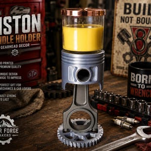 May include: A silver-colored engine piston and connecting rod assembly mounted on a circular gear base, topped with a glass candle holder containing yellow wax. The assembly features a metallic finish and industrial design elements.