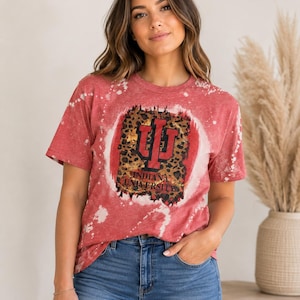 May include: A red short-sleeve t-shirt with a bleached splatter pattern. The center features a rectangular graphic with a leopard print background, a red Indiana University logo, and the text Indiana University in black capital letters.