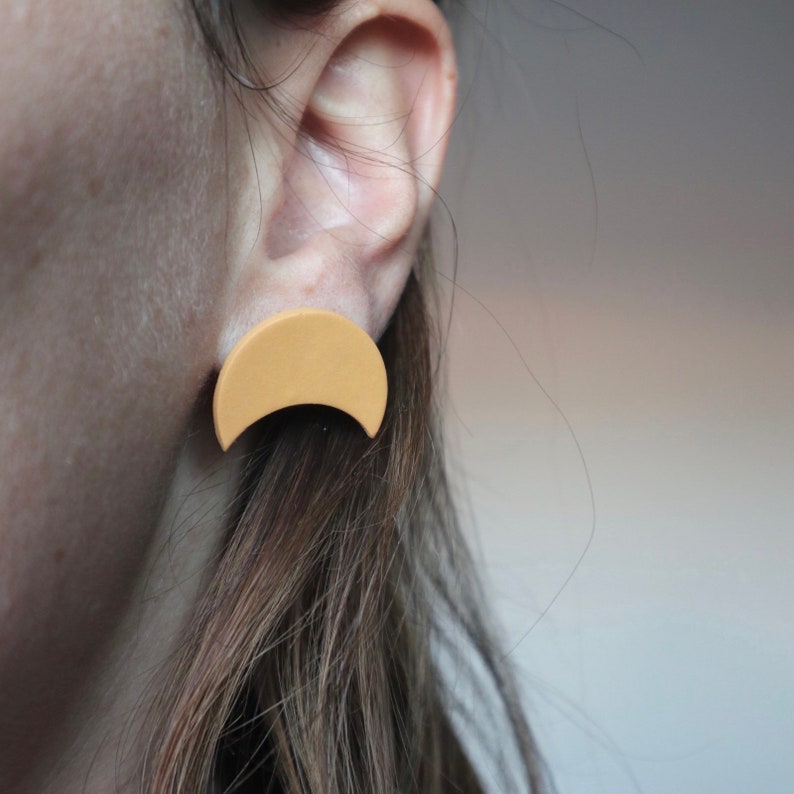May include: A close-up of an ear with a light yellow crescent-shaped stud earring. The earring has a matte finish and a simple, modern design. The person has long, brown hair. The earrings are a warm, neutral colour.