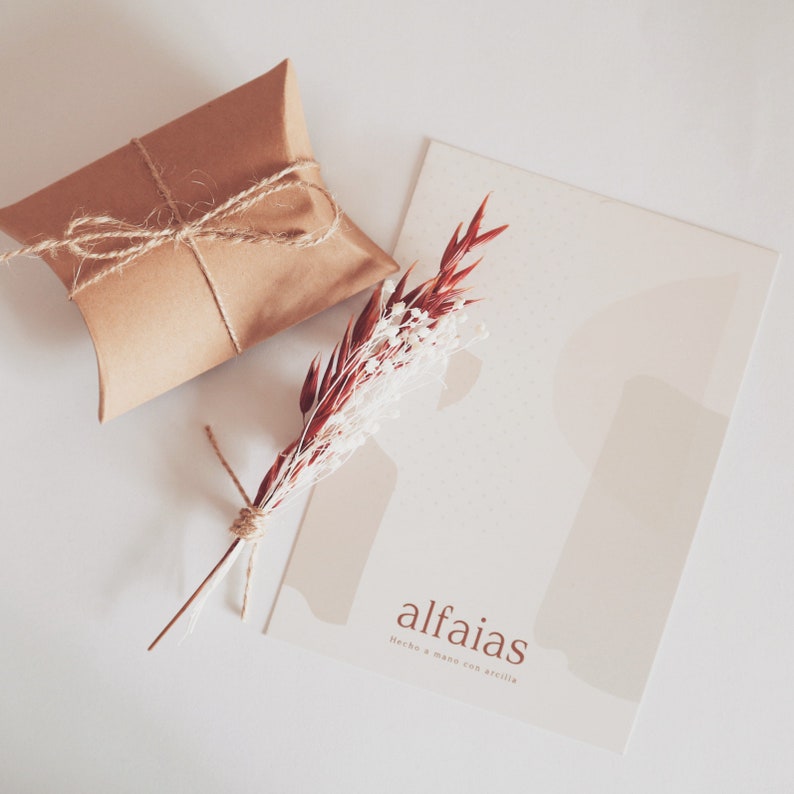 May include: A brown gift box tied with twine, a sprig of dried red and white flowers, and a card with the word "alfaias" in a serif font. The card has a neutral colour palette with abstract shapes.