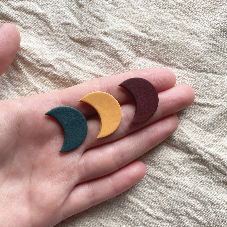 May include: Three crescent moon-shaped objects in teal, yellow, and burgundy are displayed. The objects are held in a person's hand against a neutral background. The moons are likely decorative or craft elements.