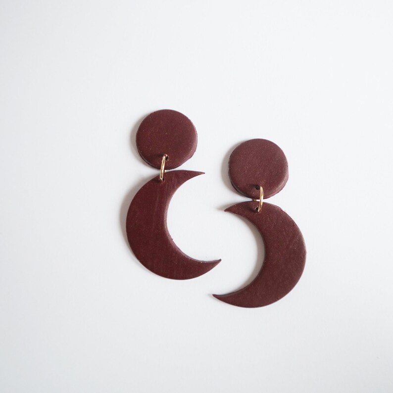 Puede incluir: Un par de pendientes de color burdeos. Cada pendiente presenta un c&iacute;rculo conectado a una forma de media luna. Los pendientes est&aacute;n suspendidos por peque&ntilde;os aros dorados. El fondo es blanco.