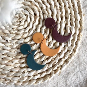 Puede incluir: Tres pares de pendientes hechos a mano de arcilla polim&eacute;rica con tem&aacute;tica celestial. Los pendientes presentan una combinaci&oacute;n de c&iacute;rculos y medias lunas en tonos verde azulado, amarillo mostaza y burdeos. Cada pendiente est&aacute; suspendido de un alambre dorado.