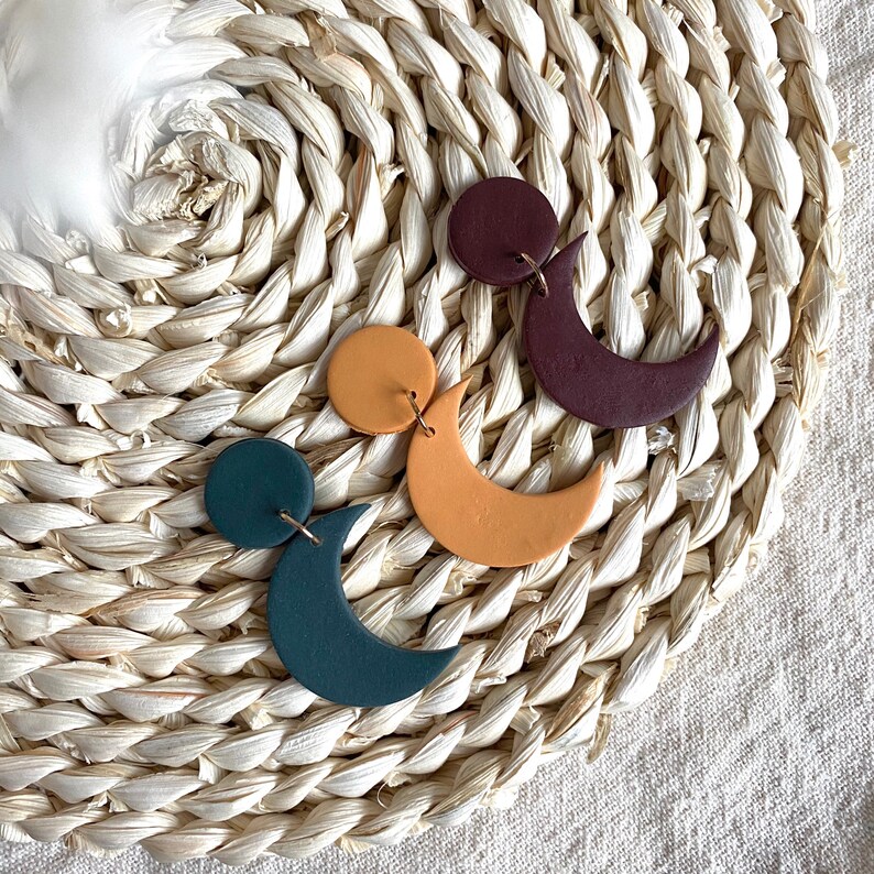 Puede incluir: Tres pares de pendientes hechos a mano de arcilla polim&eacute;rica con tem&aacute;tica celestial. Los pendientes presentan una combinaci&oacute;n de c&iacute;rculos y medias lunas en tonos verde azulado, amarillo mostaza y burdeos. Cada pendiente est&aacute; suspendido de un alambre dorado.