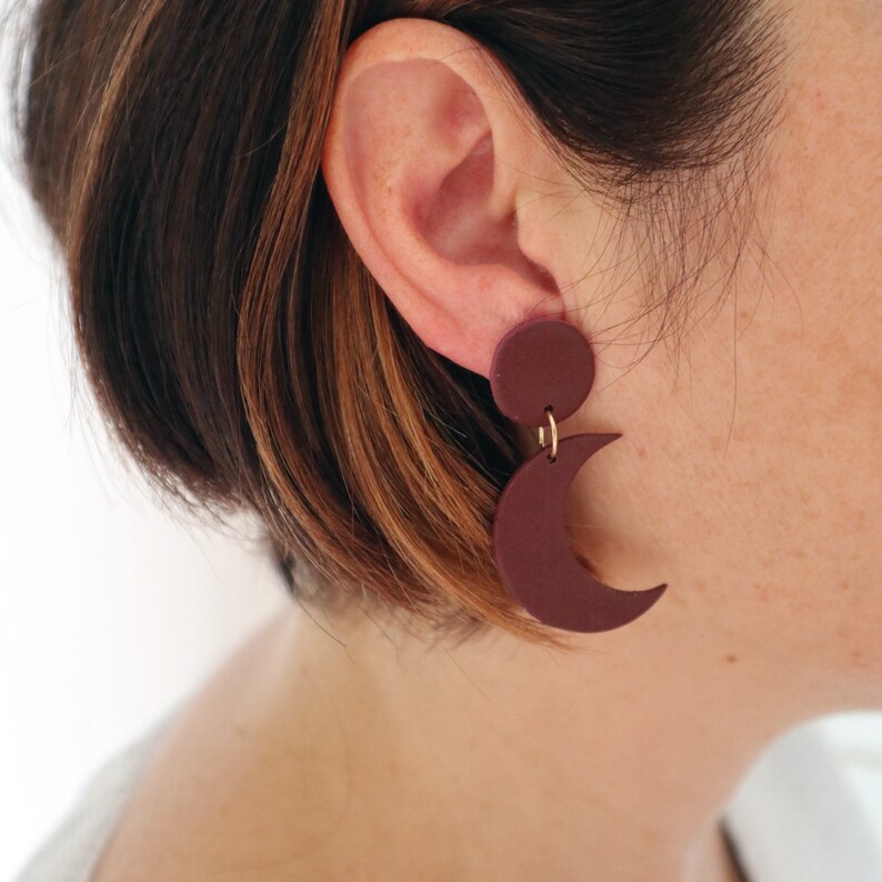 Puede incluir: Pendientes de color burdeos con una parte superior circular y una forma de media luna colgando. Los pendientes est&aacute;n conectados con un peque&ntilde;o aro dorado. Los pendientes se llevan en la oreja.