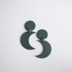 Puede incluir: Un par de pendientes de media luna verde oscuro con una parte superior circular. Los pendientes est&aacute;n conectados con peque&ntilde;os aros dorados. Los pendientes se muestran sobre un fondo blanco.