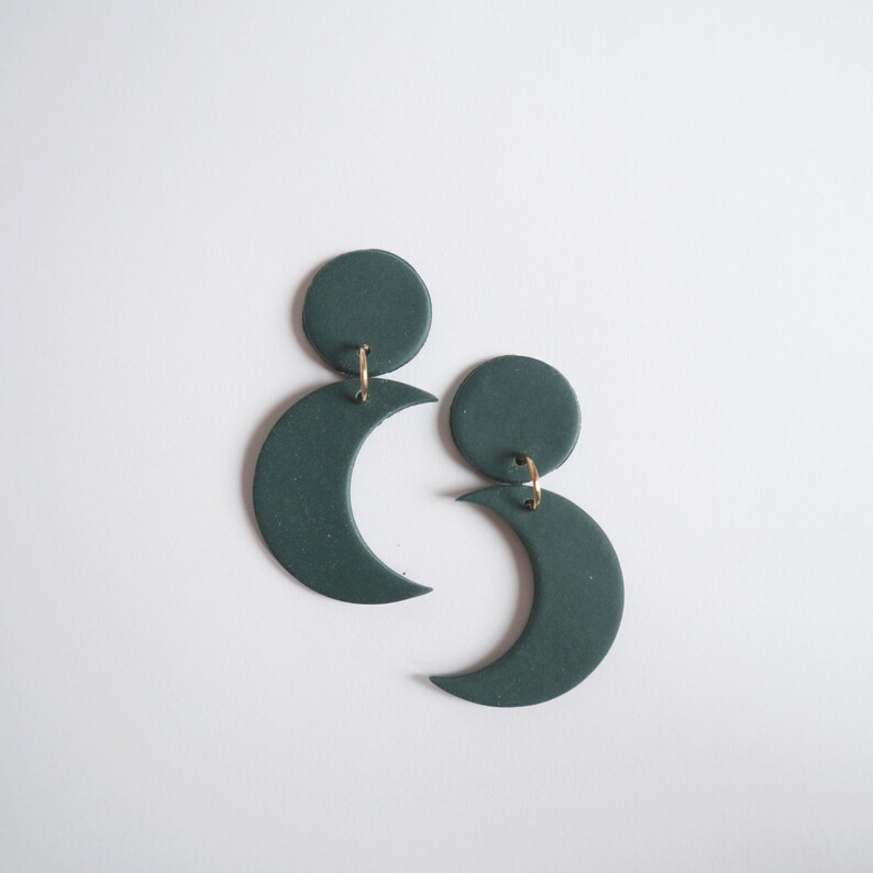 Puede incluir: Un par de pendientes de media luna verde oscuro con una parte superior circular. Los pendientes est&aacute;n conectados con peque&ntilde;os aros dorados. Los pendientes se muestran sobre un fondo blanco.