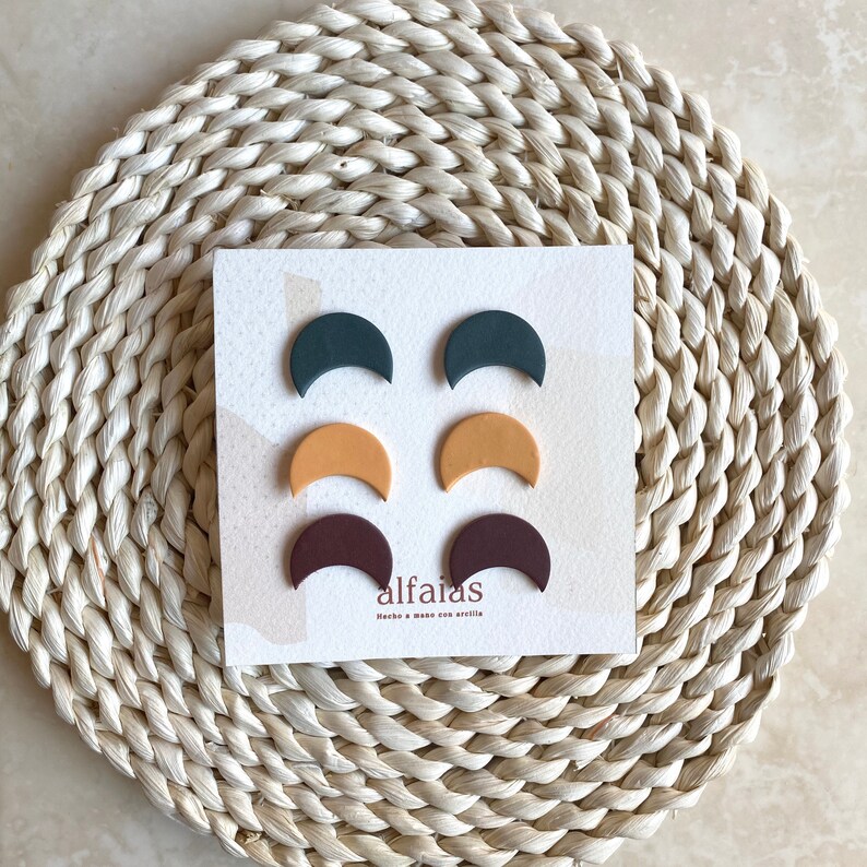 May include: Six crescent-shaped earrings in teal, mustard, and burgundy, presented on a white card. The earrings are arranged on a woven mat. The card features the brand name "alfaias".