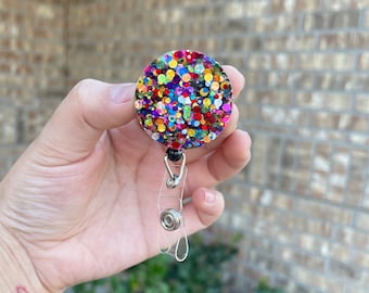 Puede incluir: Un portatarjetas redondo con un diseño de purpurina colorida. El portatarjetas tiene un cordón retráctil de plástico transparente y un clip metálico. La purpurina es una mezcla de rojo, amarillo, azul, verde y morado. El portatarjetas se sostiene en una mano.