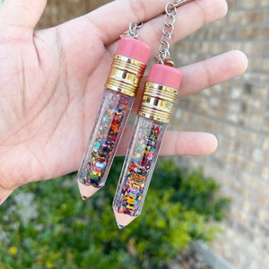 Puede incluir: Dos llaveros con forma de lápiz. La parte superior de cada lápiz es rosa, con bandas doradas. El cuerpo transparente está lleno de purpurina colorida. Cada llavero tiene una cadena y un anillo plateados.