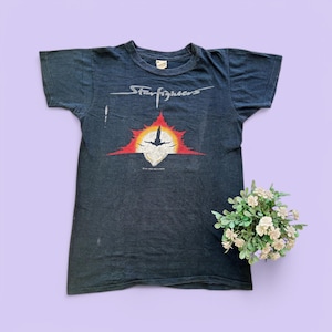 May include: Black vintage t-shirt with a graphic of a starfighter jet flying through a red and orange sun. The text "Starfighters" is printed above the graphic.
