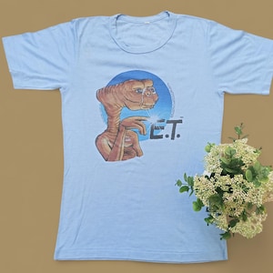 May include: Light blue t-shirt with a graphic print of E.T. the Extra-Terrestrial. The graphic shows E.T. with his finger extended, glowing with light, and the text "E.T." below.