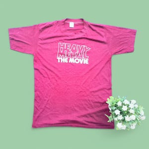May include: A maroon-colored t-shirt with the text "HEAVY METAL THE MOVIE" in white block letters. The shirt is laid flat, with short sleeves and a crew neck. A small bouquet of white flowers sits to the side.