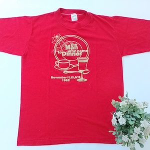 Peut inclure: T-shirt rouge avec un motif doré représentant une toile d'araignée, deux verres et une assiette avec une tasse. Le texte sur le motif indique "The Man Who Came to Dinner" et "November 11, 12, & 13, 1982".