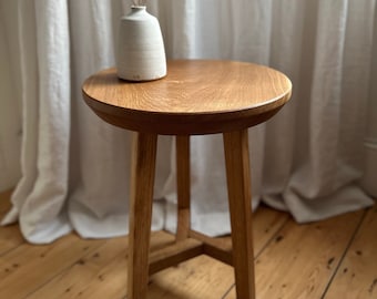 May include: A round wooden side table with a simple design. The table has a light wood finish and three legs. A white ceramic vase with dried flowers sits on top of the table.