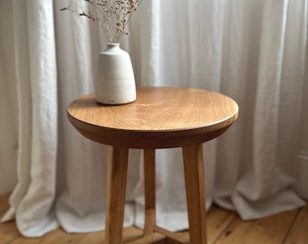 Può includere: Un tavolino rotondo in legno con tre gambe dritte e un supporto centrale. Un piccolo vaso in ceramica bianco sporco contenente rametti di fiori secchi è appoggiato sulla superficie in legno marrone chiaro del piano del tavolo.