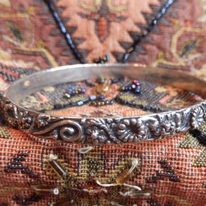 May include: A silver-toned metal bangle bracelet featuring a continuous embossed pattern of swirling scrolls, small floral motifs, and textured circular elements. The surface of the bracelet has a polished finish with darkened recessed areas to highlight the details.