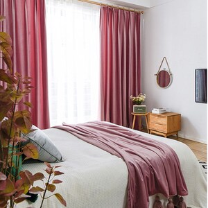 May include: A bedroom with a white bed with a pink blanket and two pink velvet curtains. There is a small wooden side table with a lamp and a mirror on the wall.