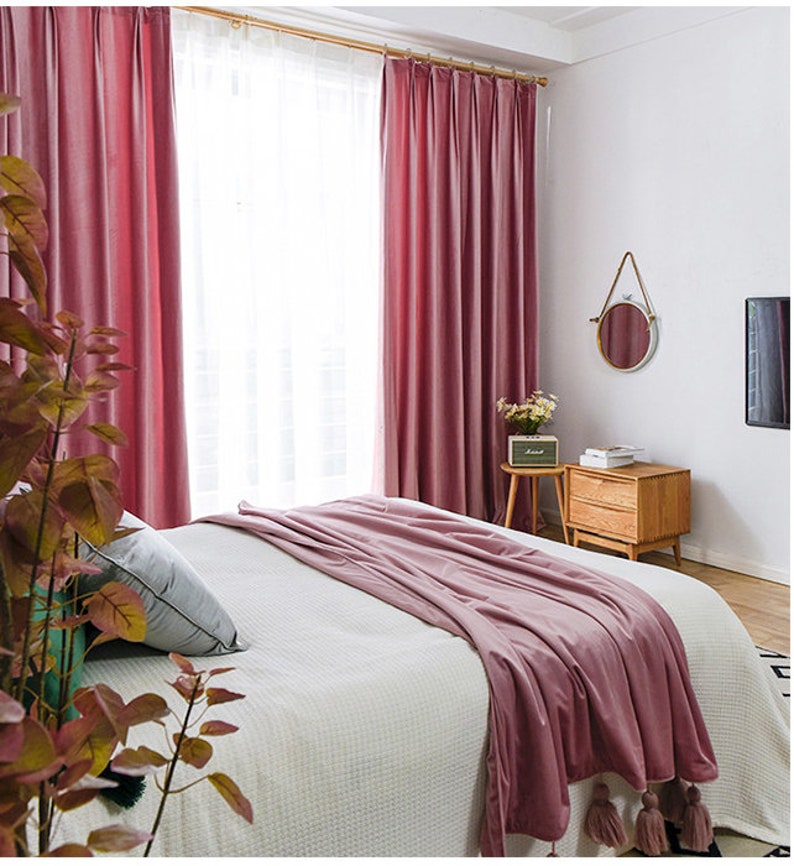 May include: A bedroom with a white bed with a pink blanket and two pink velvet curtains. There is a small wooden side table with a lamp and a mirror on the wall.