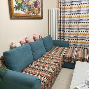 May include: A teal blue sectional sofa with a patterned throw blanket in shades of orange, black, and white. The sofa is in a living room with a white radiator, a framed picture on the wall, and a small table with a white surface.