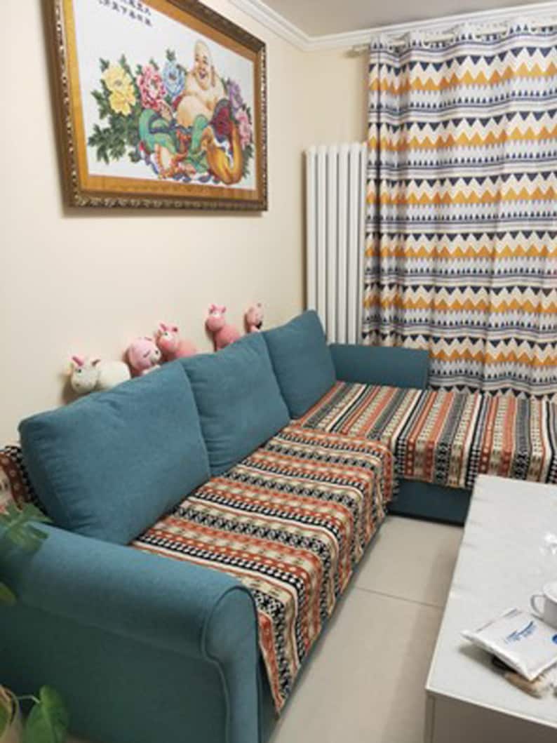 May include: A teal blue sectional sofa with a patterned throw blanket in shades of orange, black, and white. The sofa is in a living room with a white radiator, a framed picture on the wall, and a small table with a white surface.