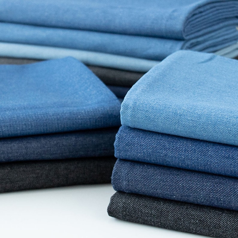 Op de afbeelding: Een stapel denim stof in verschillende tinten blauw, van licht tot donker, met een paar zwarte stukken aan de onderkant.