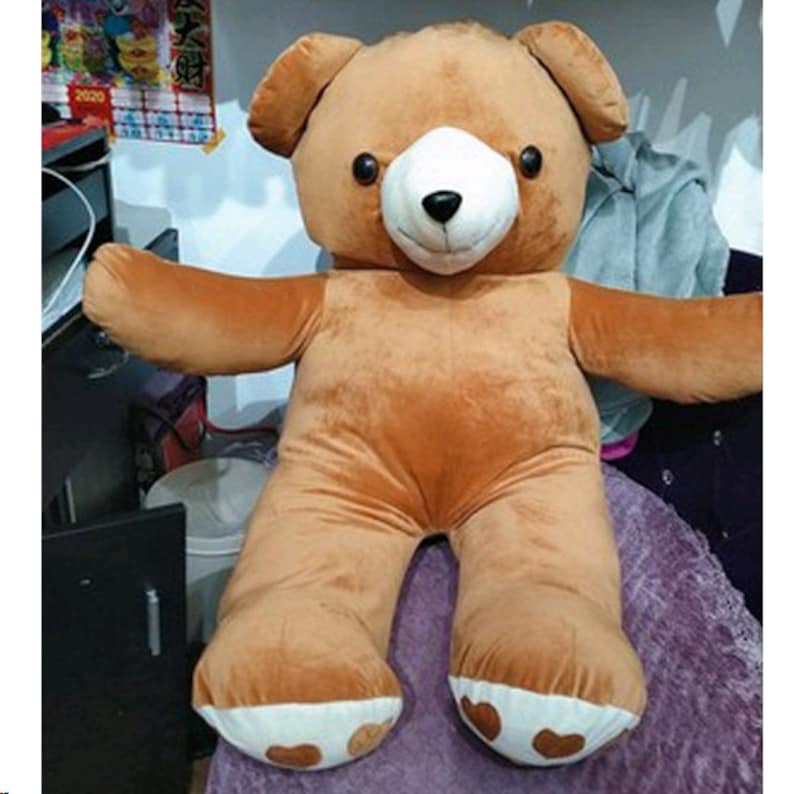 May include: A large brown teddy bear with white paws and a white face. The bear is sitting on a purple and white blanket.