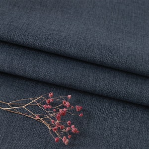 May include: A dark charcoal grey fabric with a visible woven texture and a folded edge. A small, dried sprig of delicate pink baby's breath flowers with thin, light brown stems rests on the surface of the fabric, adding a natural contrast.