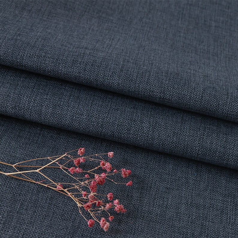 May include: A dark charcoal grey fabric with a visible woven texture and a folded edge. A small, dried sprig of delicate pink baby's breath flowers with thin, light brown stems rests on the surface of the fabric, adding a natural contrast.