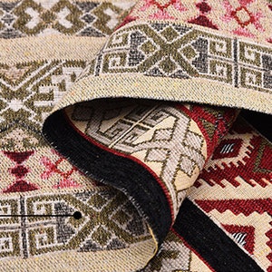 May include: Close-up of a beige and brown woven rug with geometric patterns in shades of red, green, and black.