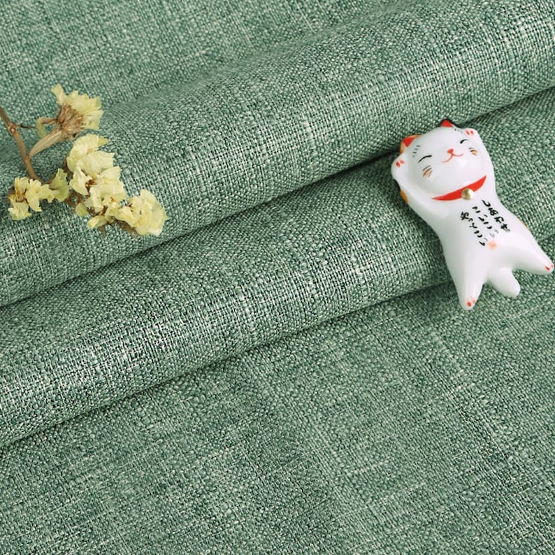 May include: A sage green fabric with a visible woven texture, featuring a small white ceramic cat figurine with a red collar and black Japanese text on its body, accompanied by a small sprig of dried yellow flowers resting on the folded material.